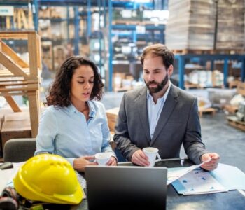 Two business people checking list and inventory on laptop. Businesswoman sharing the stock details with warehouse manager. Both standing at a table with a cup of coffee.